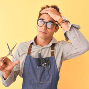 Handsome hairdresser man wearing apron holding scissors over isolated yellow background stressed with hand on head, shocked with shame and surprise face, angry and frustrated. Fear and upset for mistake.
